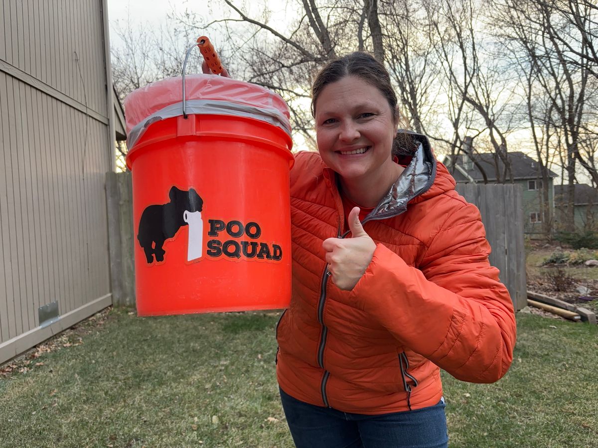 Smiling Poo Squad crew member in an orange jacket gives a thumbs up while holding an orange Poo Squad bucket in a yard