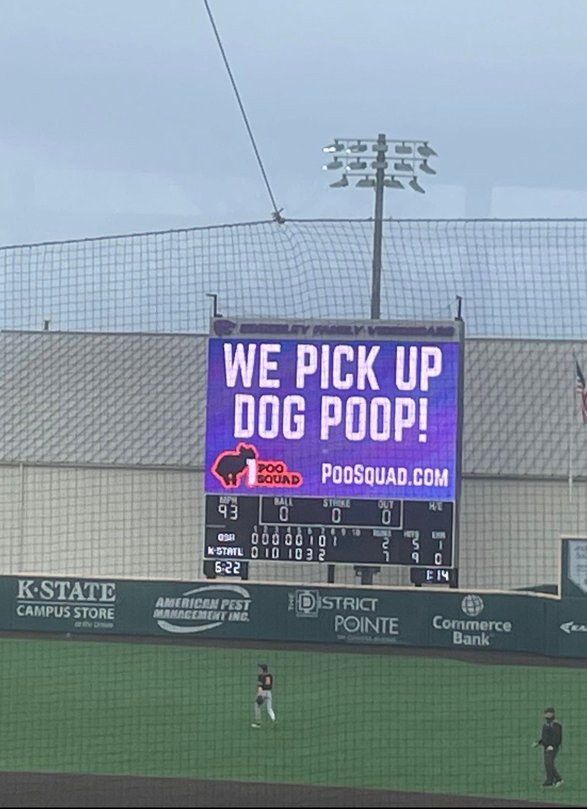 K-State baseball stadium video board showing a Poo Squad ad that says We Pick Up Dog Poop! and PooSquad.com