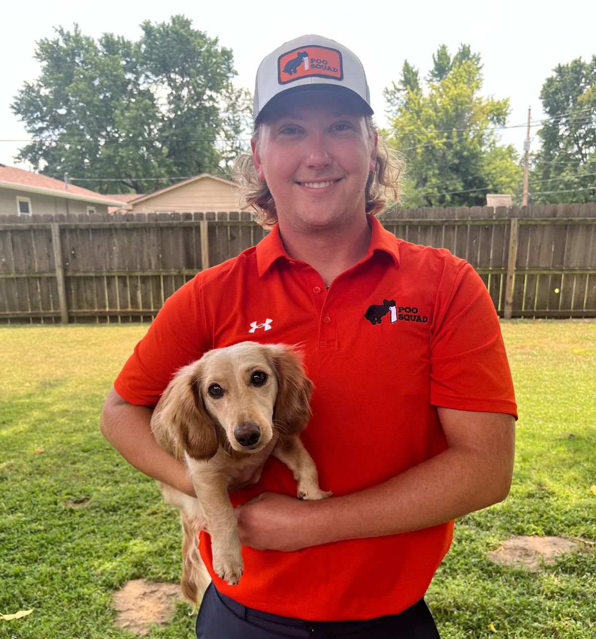 Smiling Poo Squad crew member in orange polo and cap holding a cream long-haired dachshund in a grassy backyard