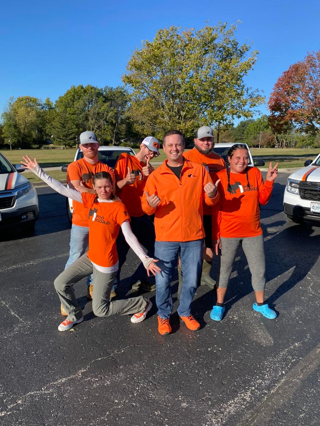 Poo Squad crew in orange shirts posing cheerfully in a parking lot beside white company trucks on a sunny fall day