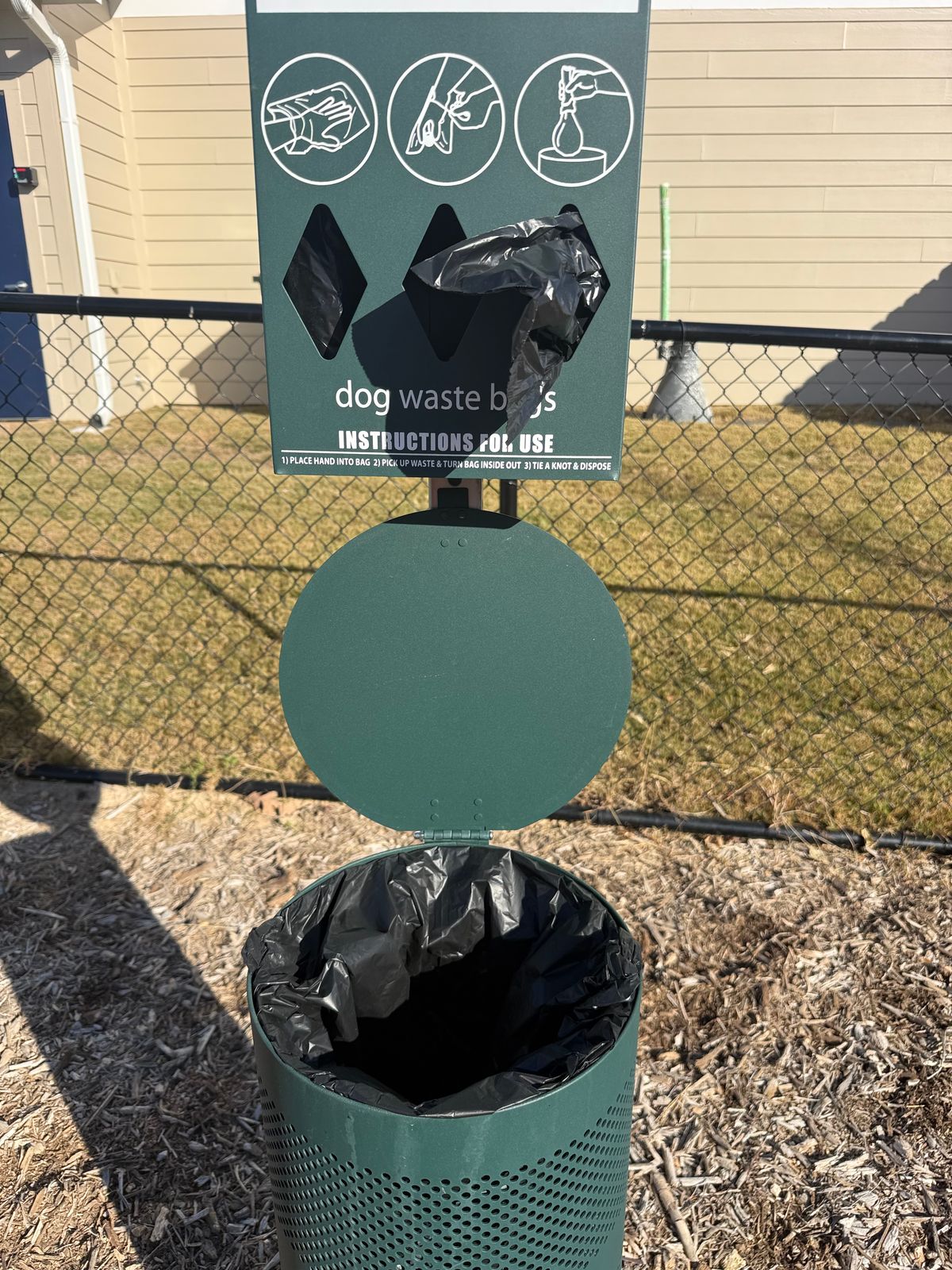 Green pet waste station with bag dispenser and freshly lined trash bin beside a fenced grassy area