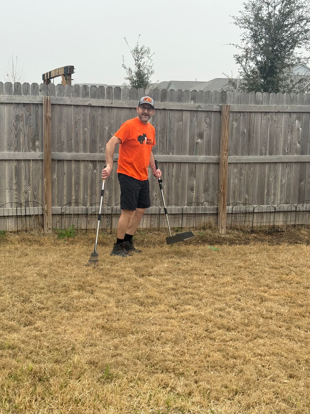 Poo Squad crew member in orange shirt holding a rake and scooper in a fenced backyard with dry grass