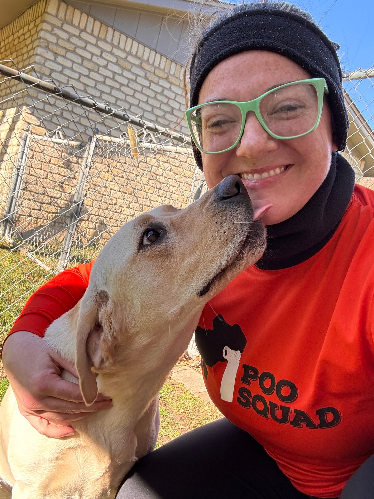 Poo Squad crew member smiling as a yellow Labrador licks her chin in a sunny fenced backyard